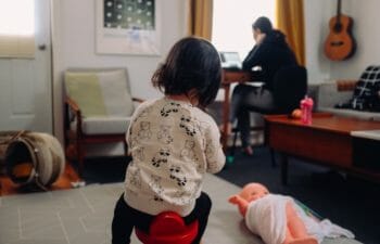 Child plays in the foreground, while the child's mom works on a laptop in the background.