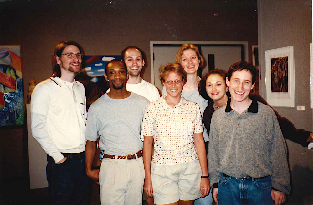 Photo of Oen among a group of seven students, in a dim gallery space with paintings, photographs, and graphic prints on the walls behind them.