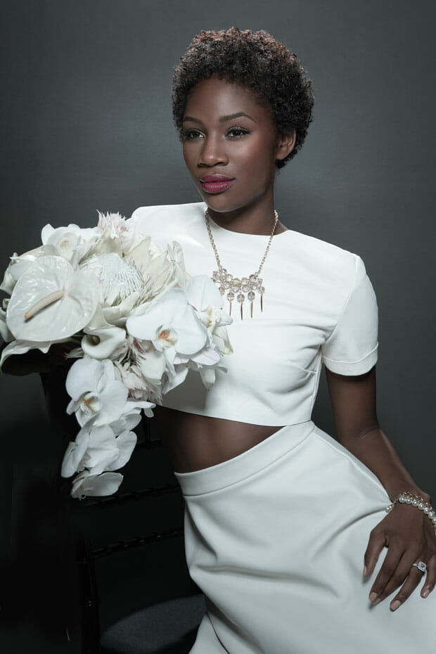 A young model with short, dense curls and crisp makeup poses in a white crop-top and white skirt by a vase of glassy orchids against a neutral background.