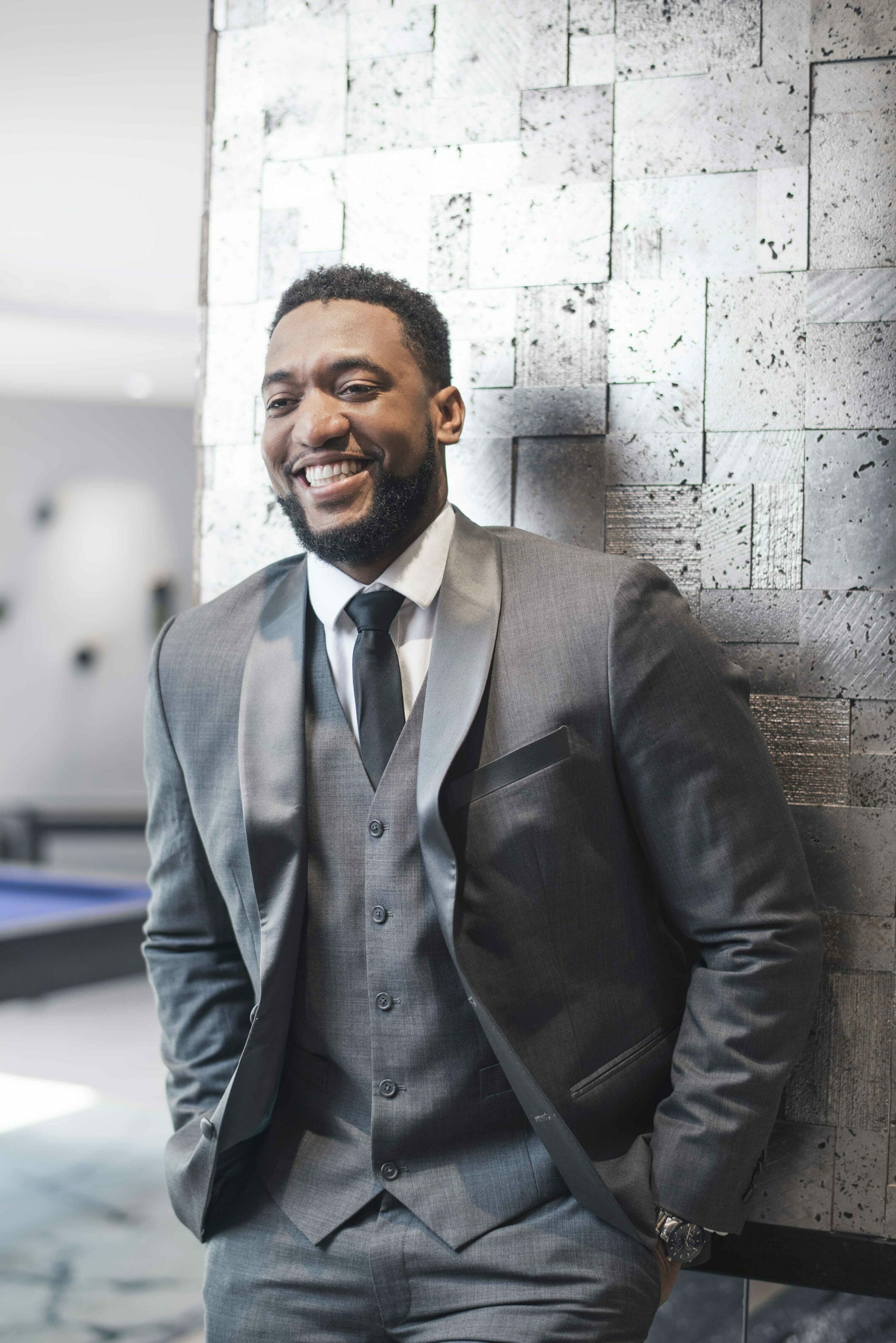 Man in a grey, tuxedo, smiling and posing against a textured background.