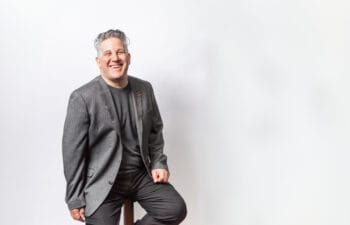 Man with silver hair smiling and waring a gray suit sitting on a stool