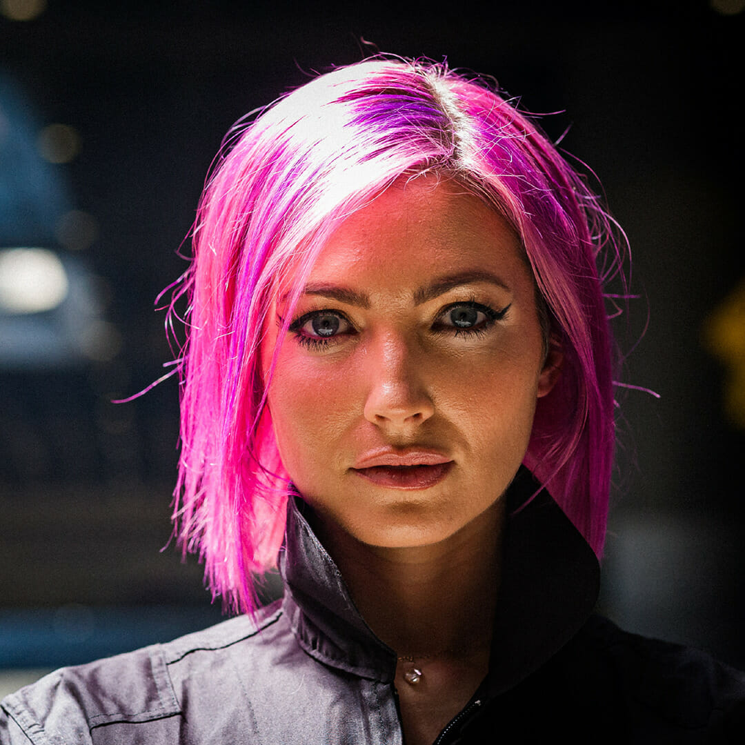 Headshot of MegZany with pink hair against a black brick wall.