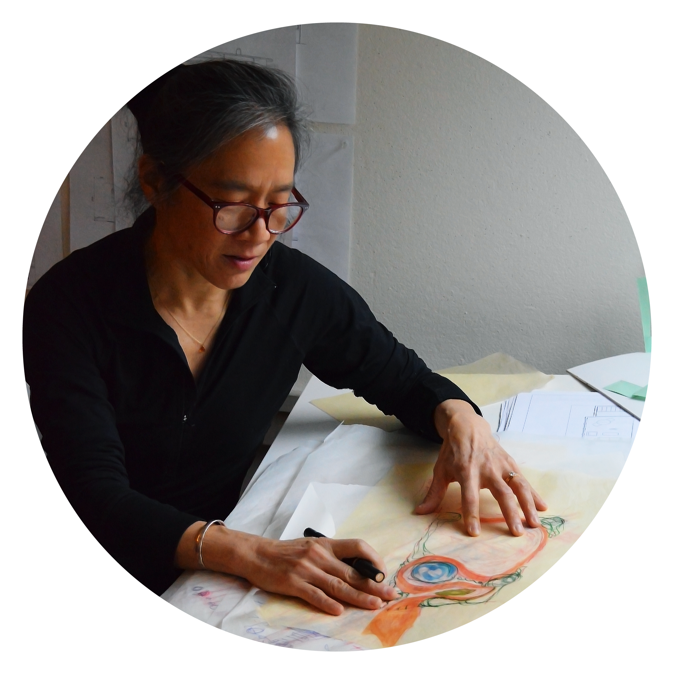 Suenn leaning over a table with scattered papers, with a pen in her hand. Suenne is wearing a black outfit and reading glasses rest on her face.