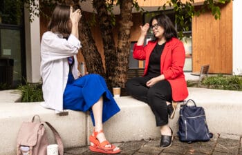 Two women facing each other on a curved white bench while they practice American Sign Language. Text says: Deaf-friendly curved seating makes signing easier, and points to the bench. A label "Sensory retreat space" points to a lone bench in the background. And it says "BONUS: Sturdy, fat-friendly benches" as well.