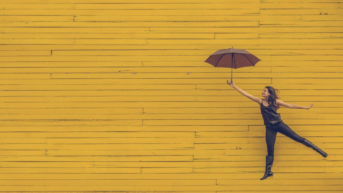 Woman holding against a yellow background holding an umbrella as if she's flying