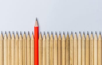 A row of similar-looking pencils with one red pencil that is pushed higher up than the rest.