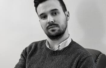 Black and white photographic portrait of a white man with dark hear wearing a collared shirt and sweater looking into the camera.