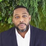 Williams,Jabali_Headshot A portrait of a black man wearing a blue blazer in front of a plant backdrop.