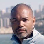 Black man wearing a light grey shirt in front of a city skyline