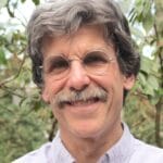 White man with grey hair, glasses, and a mustache in front of a leafy background