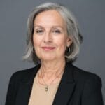 Grey-haired, white woman wearing a black blazer, tan shirt, and a silver necklace.
