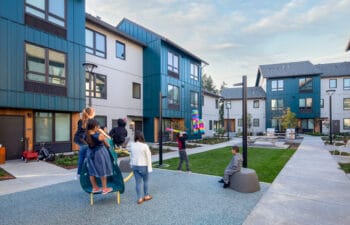 A group of children in a playspace look on at a blindfolded boy swinging a baton at a piñata.