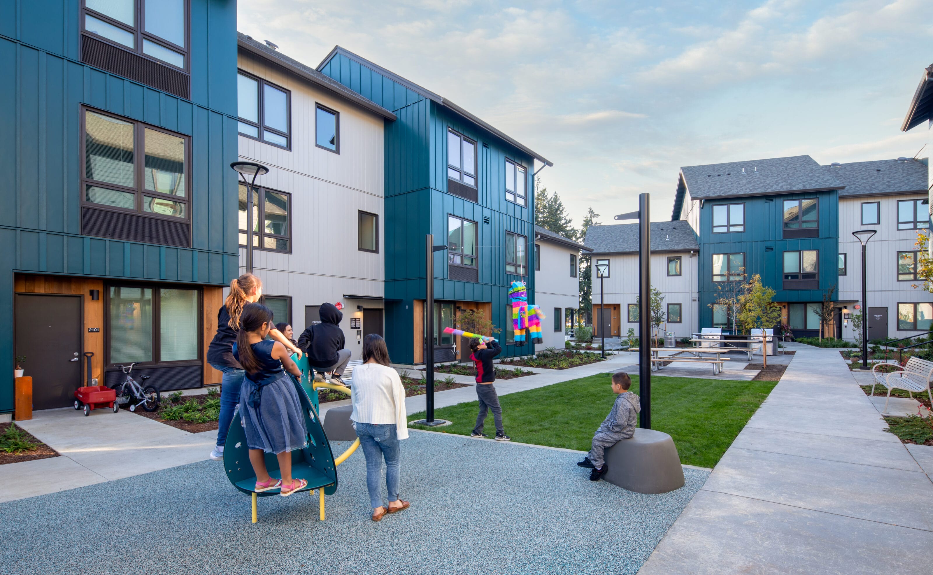 A group of children in a playspace look on at a blindfolded boy swinging a baton at a piñata.