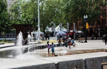 Young children play outside in a public fountain at the center of a city park while adults look onward from nearby benches.