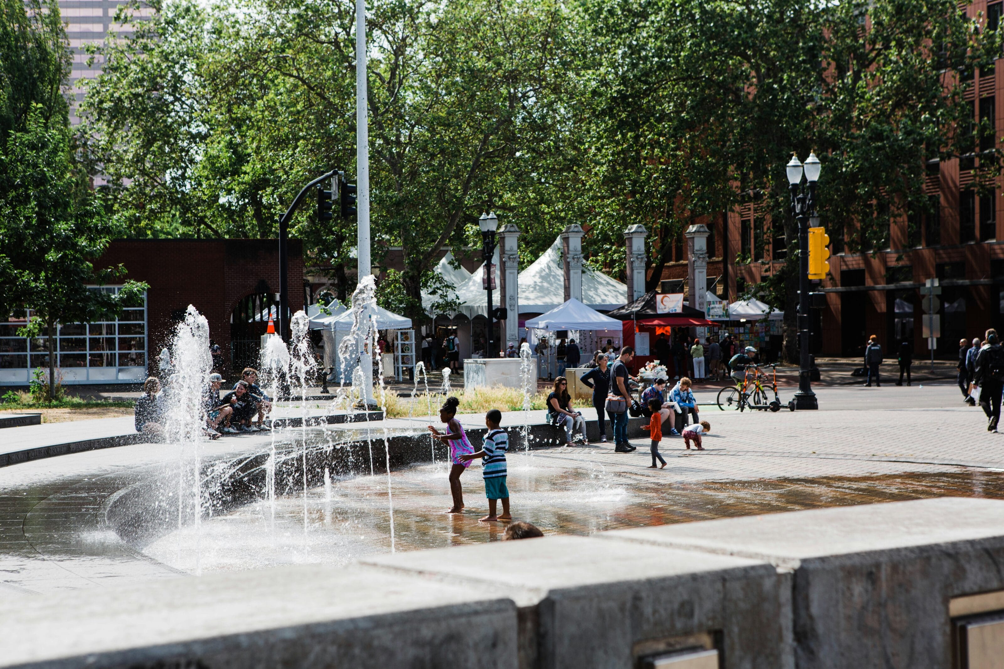 Young children play outside in a public fountain at the center of a city park while adults look onward from nearby benches.