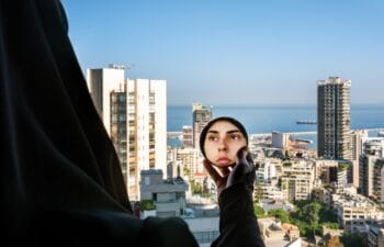 A photograph by Rania Matar titled "Alae (In the Mirror), Beirut, Lebanon, 2020." A figure wrapped in dark fabric stands on a high-rise balcony looking out over the densely packed buildings of Beirut toward the Mediterranean Sea. The person holds up a circular hand mirror in front of the skyline, revealing a clear reflection of their face looking directly into the lens.