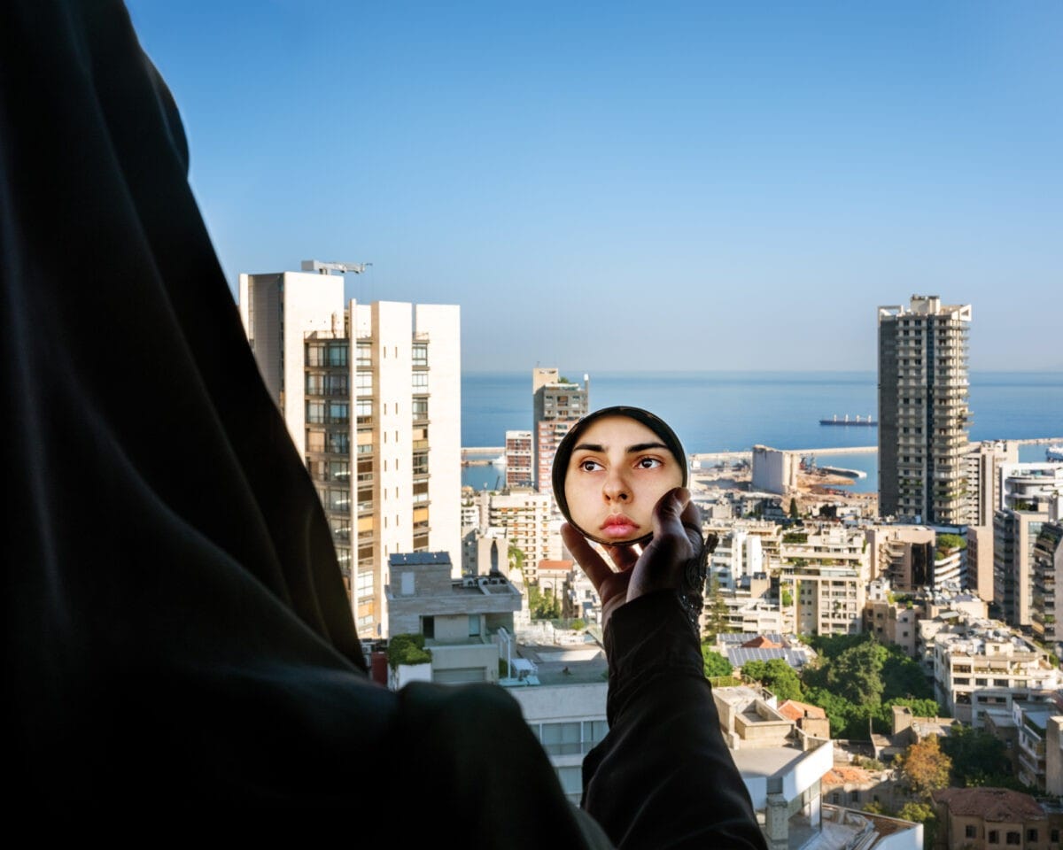RM-046_SHE-e1618607319229 A photograph by Rania Matar titled "Alae (In the Mirror), Beirut, Lebanon, 2020." A figure wrapped in dark fabric stands on a high-rise balcony looking out over the densely packed buildings of Beirut toward the Mediterranean Sea. The person holds up a circular hand mirror in front of the skyline, revealing a clear reflection of their face looking directly into the lens.