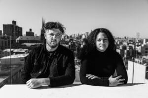 A black and white outdoor portrait featuring Ryan Swanson and Maeghann Coleman of The Urban Conga, seated next to each other and smiling at the camera against a soft-focus background.