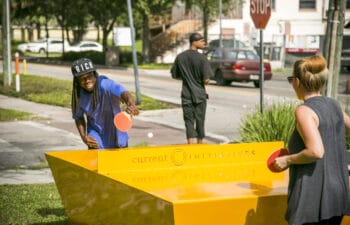 An interactive public installation featuring a ping pong table setup on an urban sidewalk, with two people actively playing alongside a large yellow structure and city street scenery in the background.