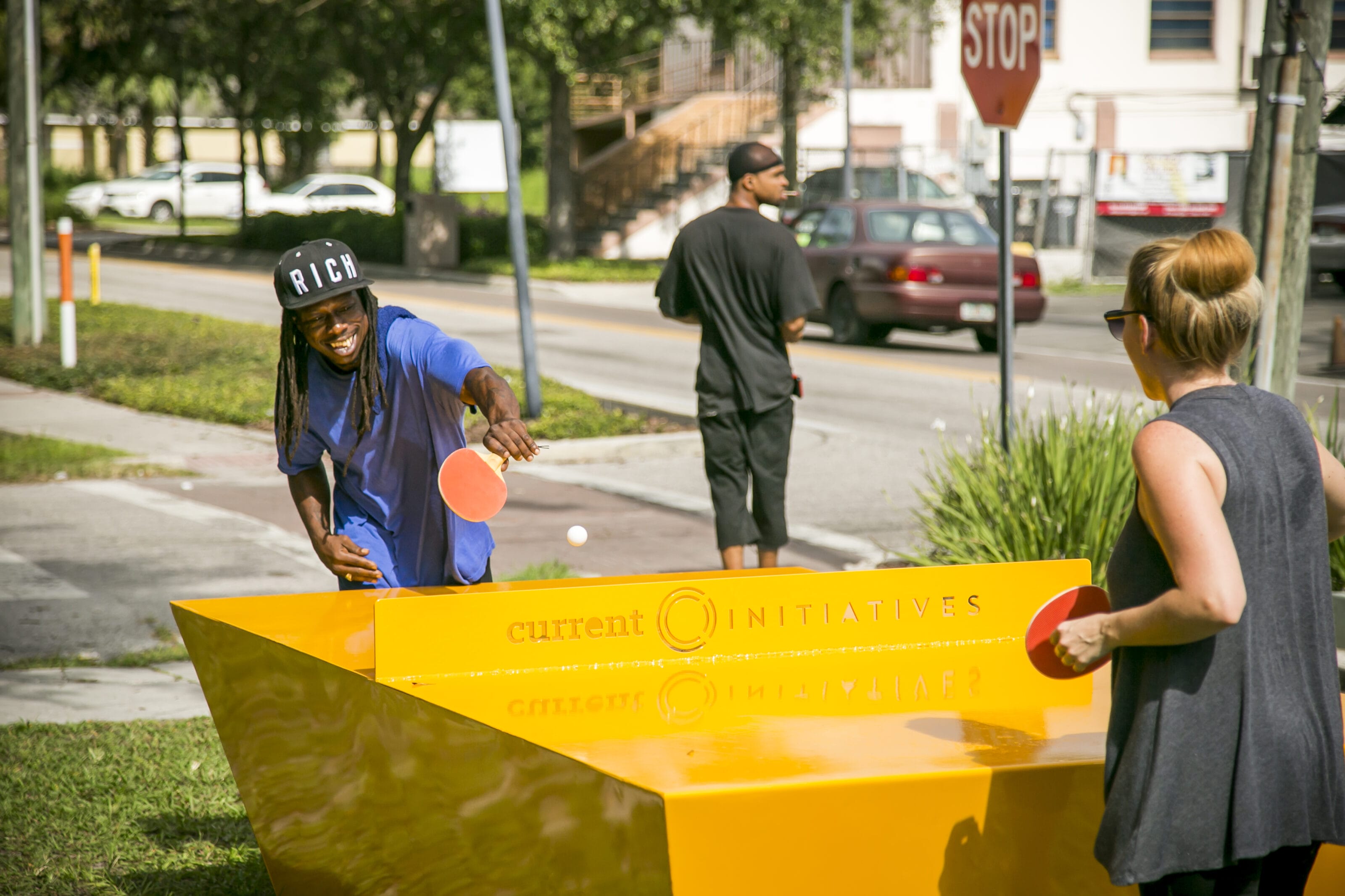 An interactive public installation featuring a ping pong table setup on an urban sidewalk, with two people actively playing alongside a large yellow structure and city street scenery in the background.